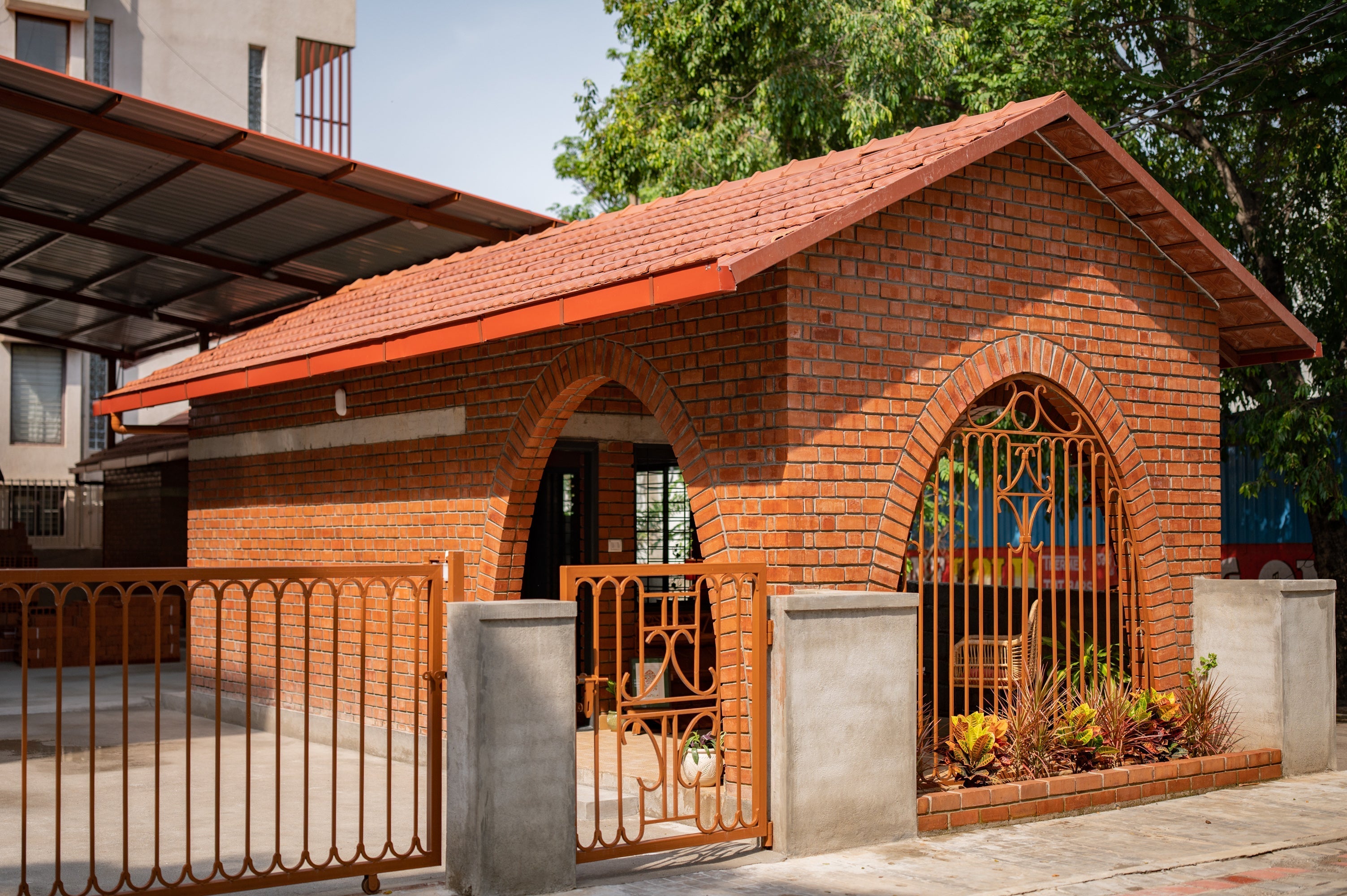 A small brick house with an arched doorway, tiled roof, and decorative metal gate. Concrete pillars and plants enhance its welcoming appearance.