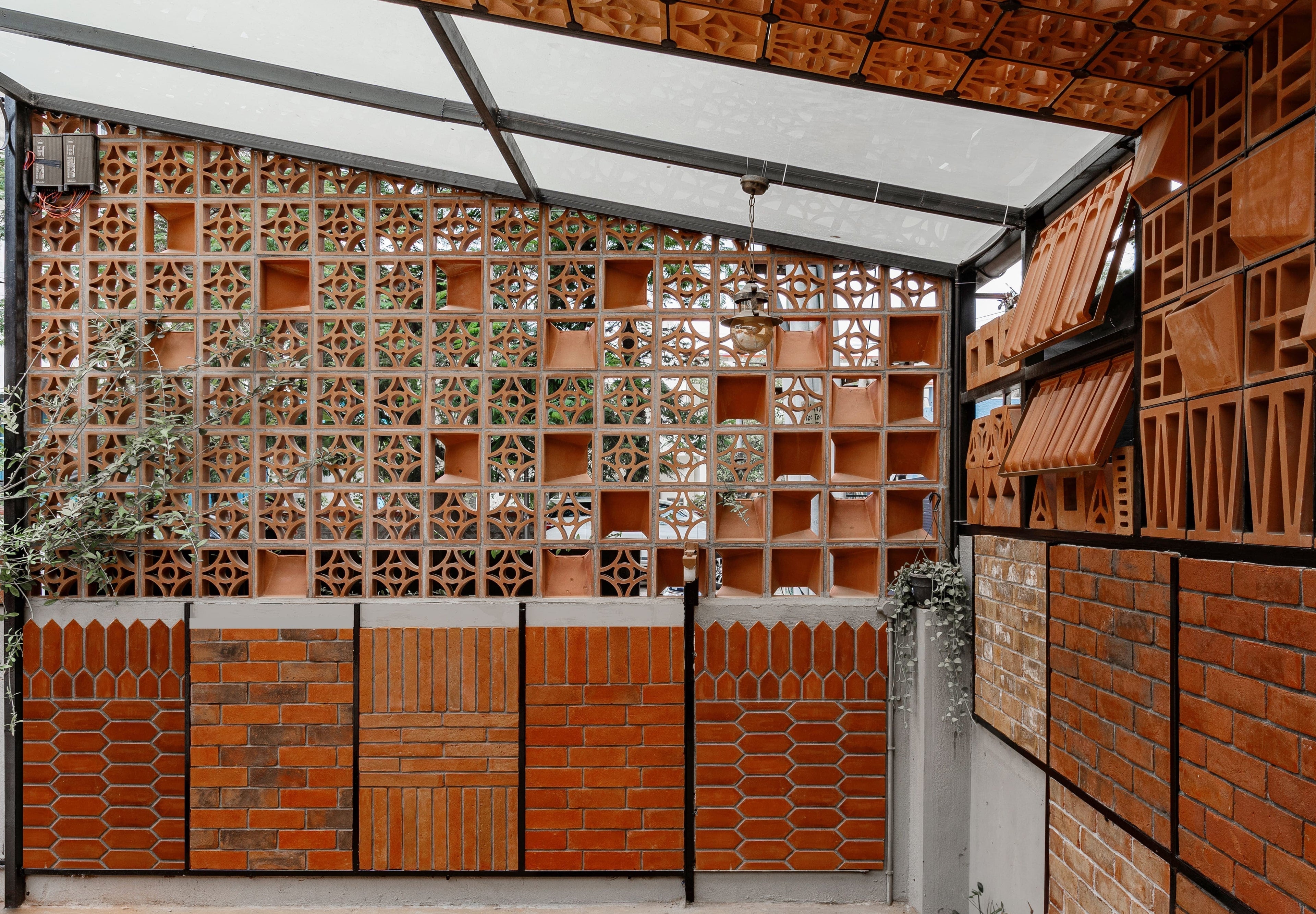 Covered outdoor space with a transparent roof, featuring various red brick patterns on walls, creating a warm, textured look. Sparse green plants add a natural touch.